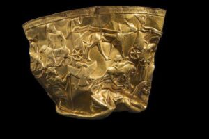 نمای نزدیک از جام زرین حسنلو با نقوش برجسته اساطیری و باستانی ایران در پس‌زمینه مشکی یکدست که جزئیات حکاکی‌های طلایی را با تضاد نوری شدید نشان می‌دهد