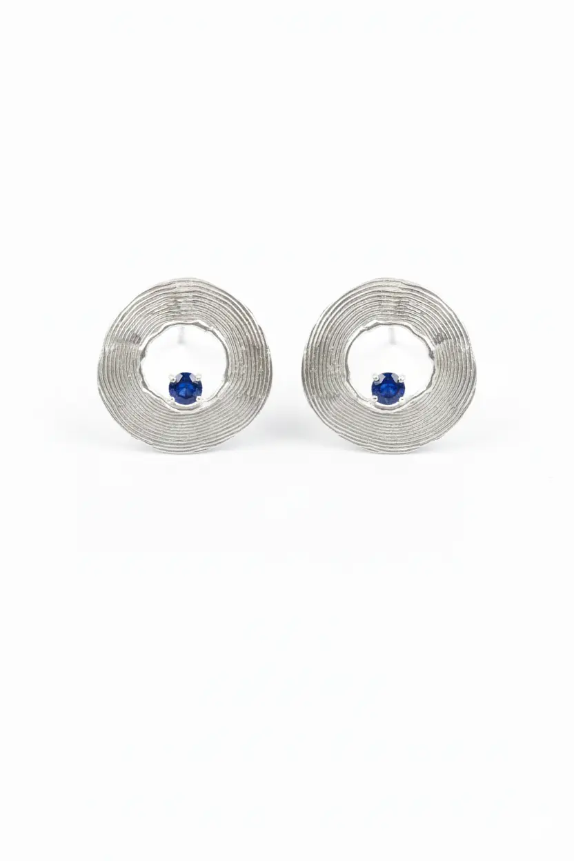 گوشواره دایره نقره گوشوارههای دایرهای نقره با بافتهای هممرکز و یک سنگ سواروسکی آبی کوچک در پایین کادر، عکاسی شده روی زمینه سفید.