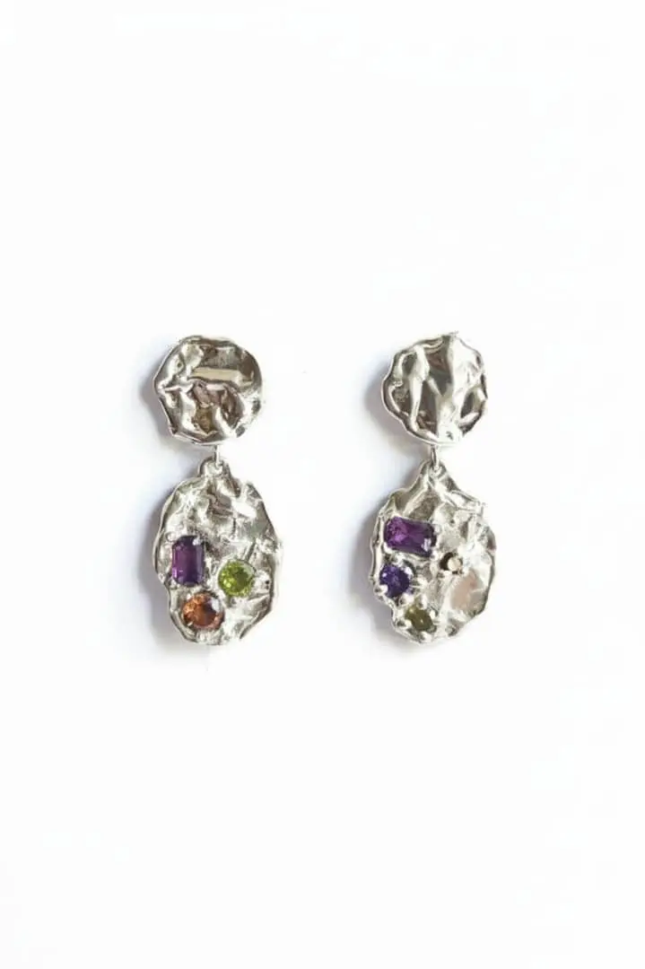 گوشواره نقره با سنگ اتمی گوشوارههای نقره دستساز با سطح بافتدار و نامنظم دارای چهار سنگ اتمی درخشان به رنگهای بنفش، سبز و نارنجی، عکاسی شده روی زمینه سفید.
