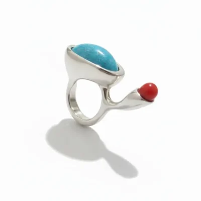 انگشتر نقره دست‌ساز با طراحی نامتقارن دارای یک سنگ فیروزه بیضی بزرگ و یک سنگ مرجان قرمز کروی، عکاسی شده روی پس‌زمینه سفید.