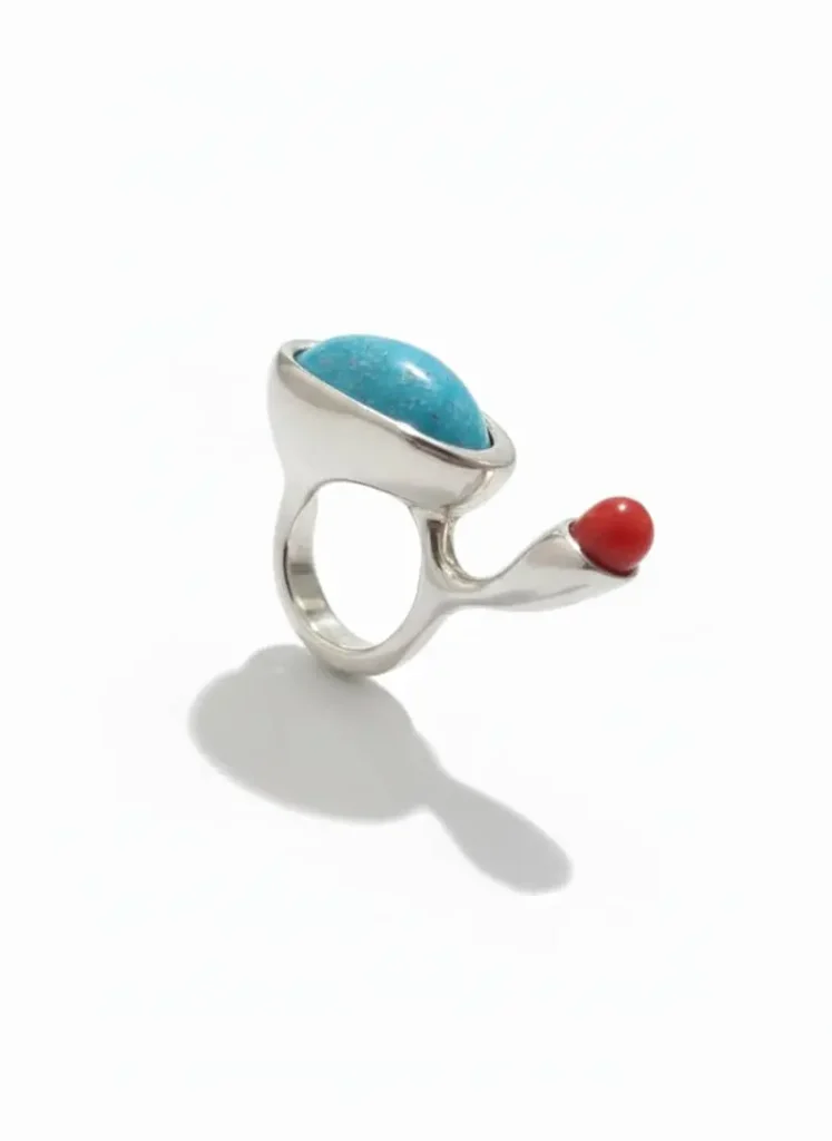 انگشتر نقره دست‌ساز با طراحی نامتقارن دارای یک سنگ فیروزه بیضی بزرگ و یک سنگ مرجان قرمز کروی، عکاسی شده روی پس‌زمینه سفید.