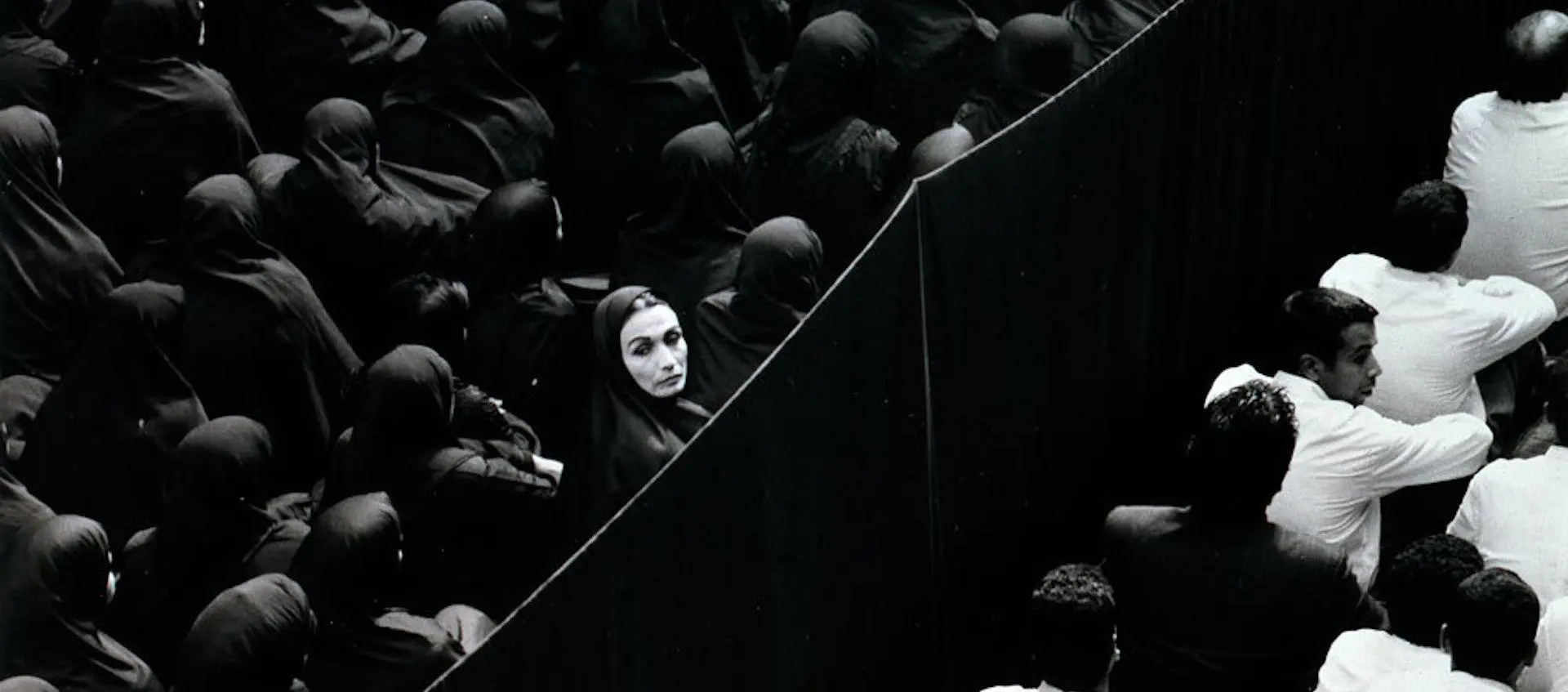 «جمعی از زنان با پوشش چادر که همگی در یک جهت قرار گرفته‌اند، در حالی که تنها یک زن در میان آن‌ها سرش را برگردانده و مستقیم به دوربین نگاه می‌کند؛ تأکیدی بر تفاوت فردی در دل یک جمع هم‌شکل.