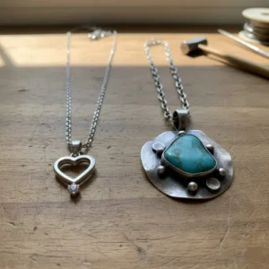 مقایسه گردنبند نقره ظریف صنعتی با طرح قلب و گردنبند دست‌ساز هنری با سنگ فیروزه درشت روی میز چوبی کارگاه