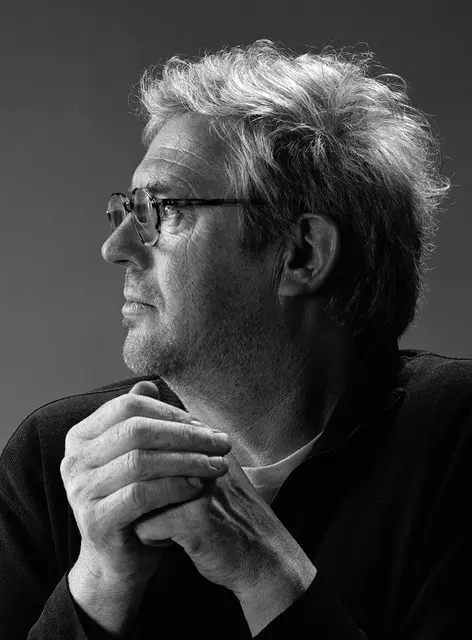 پرتره سیاه و سفید از نیمرخ مانفرد بیشوف (Manfred Bischof) با عینک، موهای جوگندمی و دستهای گرهخورده زیر چانه، مناسب برای بخش معرفی هنرمند در سایت.